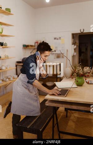 Potter talking on smart phone while using laptop on table at workshop Stock Photo