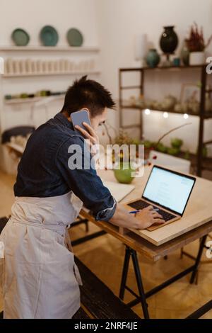 Potter with laptop talking on mobile phone at workshop Stock Photo