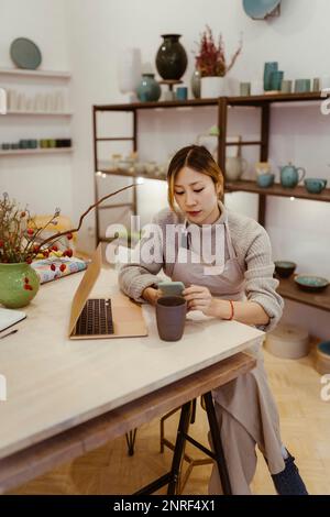 Young female entrepreneur with laptop using mobile phone while sitting at workshop Stock Photo