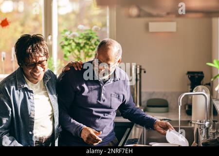 Happy senior woman with hand on shoulder of man washing dishes in kitchen at home Stock Photo