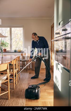 Senior man cleaning carpet with vacuum cleaner while standing at home Stock Photo