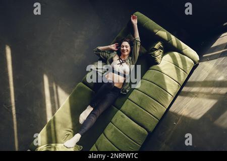 Cheerful woman in headphones lying on couch dancing to music Stock Photo