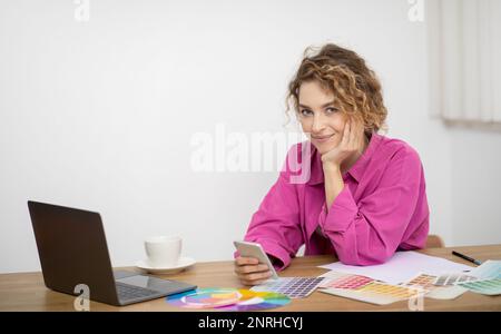 Young designer woman sitting at desk with colour swatches and using smartphone Stock Photo