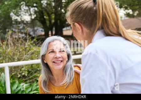 Female doctor consoling smiling senior woman in garden of nursing home Stock Photo