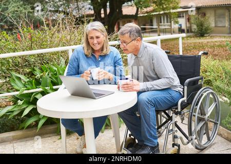 Happy senior man sitting on wheelchair discussing with woman over laptop at table in garden of nursing home Stock Photo