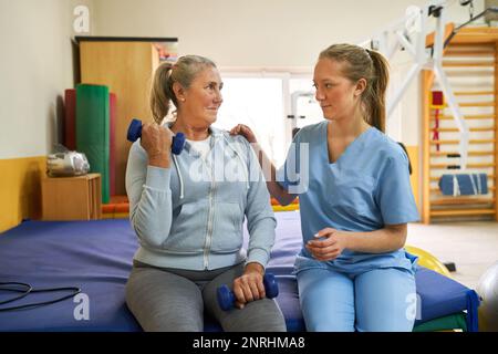 Female therapist assisting senior woman exercising with dumbbells while sitting on bed at rehabilitation center Stock Photo