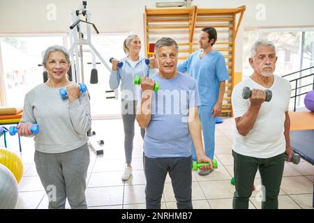 Senior men and woman exercising with dumbbells while therapist assisting elderly patient in background at nursing home Stock Photo