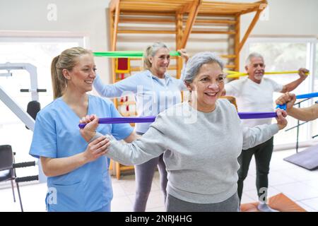 Happy young female therapist assisting elderly woman in workout with resistance band at nursing home Stock Photo
