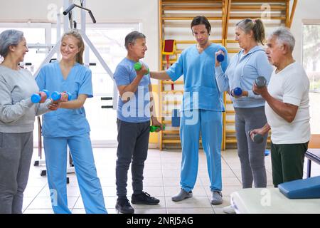 Female nurse assisting senior men and woman exercising with dumbbells at rehab center Stock Photo
