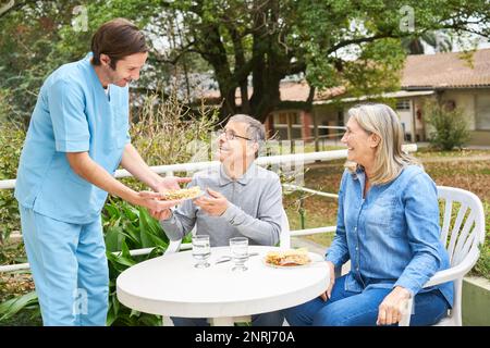 Smiling male caregiver looking at senior man and woman having meal at table in garden of nursing home Stock Photo
