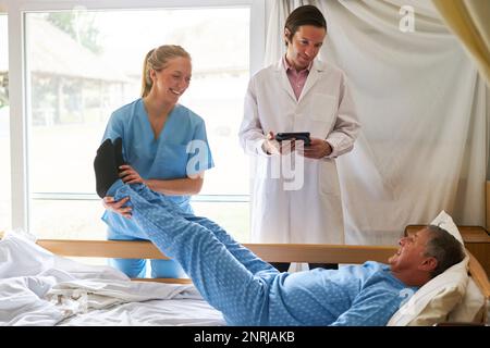 Doctor standing by smiling female nurse lifting legs of senior man lying in bed at nursing home Stock Photo