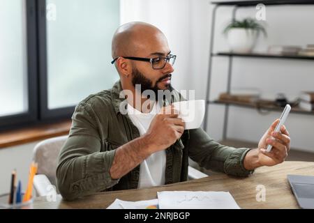 Happy latin businessman drinking coffee and using cellphone, sitting at desk at home office interior, free space Stock Photo