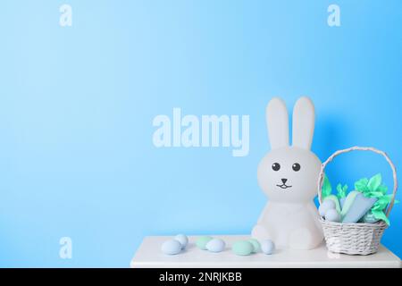 White toy Easter Bunny, basket with toy carrots and Easter eggs on blue background Stock Photo
