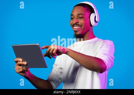 Joyful black guy using modern digital pad and wireless headphones Stock Photo