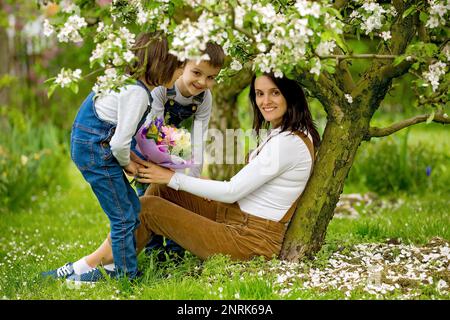 Young pregnant woman, receiving bouquet of colorful flowers from her children for Mothers Day, sitting in a beautiful spring blooming garden Stock Photo