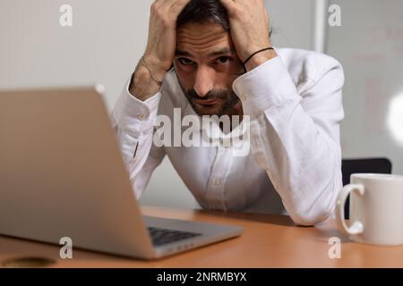 portrait of a young office worker sitting in front of his computer. He is tired and stressed and holding his head in his hands Stock Photo