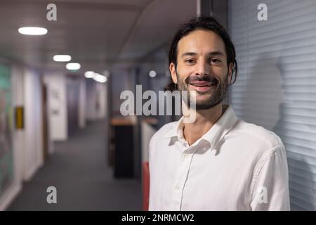 Portrait of a handsome 30-year-old man standing with his arms crossed in the hallway of his office. He is serene and smiling, happy at work. Symbol of Stock Photo