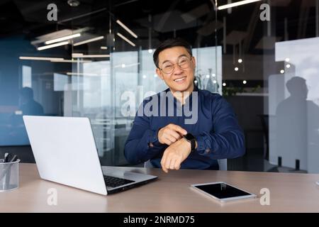 Portrait of successful smiling asian man inside office, businessman smiling and looking at camera, pointing finger at smart watch, man at workplace with laptop, programmer coding software. Stock Photo