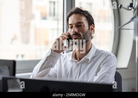 portrait of a young businessman or office worker working on his laptop in a cozy office and communicating on his mobile phone Stock Photo