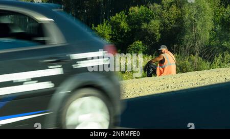 A road worker in an orange vest sits on the side of the road past passing cars while relaxing in the summer in Russia. Stock Photo