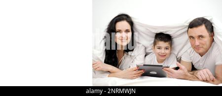 banner happy modern family finds on internet shopping sale plays games watches movies spend time together in bed under the covers three people mom dad and son look into frame smile in hands tablet Stock Photo