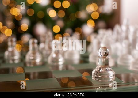 Glass-made chess figures on a chess board with a pawn in selective focus and blurred lamps in the background. Stock Photo