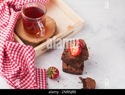 Breakfast with hot black tea, delicious homemade chocolate brownie pieces decorated with  organic strawberries. Top view, copy space, close up. Stock Photo