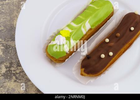 White plate with delicious green and brown glazed french dessert eclairs  on light gray cement table. Top view, close up horizontal background. Stock Photo