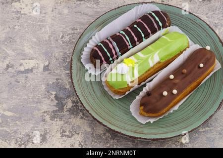 Green plate with delicious colorful green and brown glazed french dessert eclairs  on light gray cement table. Top view, close up horizontal backgroun Stock Photo