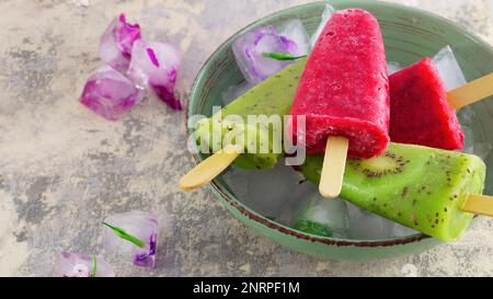 Homemade kiwi and raspberry ice cream or popsicles with frozen flowers on concrete table, frozen fruit juice, vintage style, quare. Stock Photo