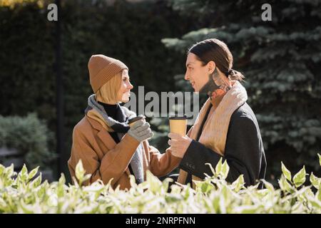 side view of cheerful woman in beige hat looking at tattooed boyfriend in coat while holding coffee to go,stock image Stock Photo