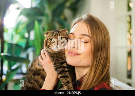 Casual Young Blonde Woman Holds Shoulder a Cute Green-eyed Scottish Tabby Cat and hugs it at Home. The concept of loving and caring for pets Stock Photo