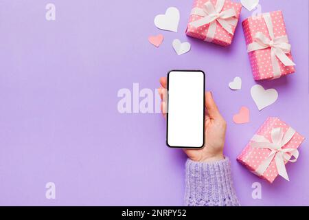 Woman hand holding mobile phone with blank screen on colored background with hearts, valentine day concept top view flat lay. Stock Photo