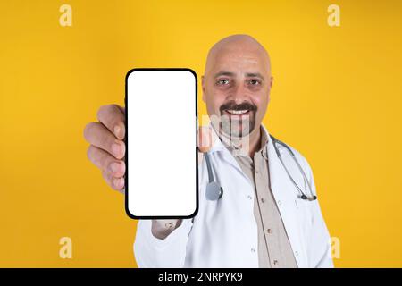 Smiling positive caucasian doctor showing mobile phone mock up. Wearing white cape and stethoscope. Recommending concept idea. Stock Photo