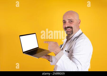 Doctor showing empty white screen of modern laptop for mock up. Online appointment or consultation concept idea. Isolated orange background. Stock Photo