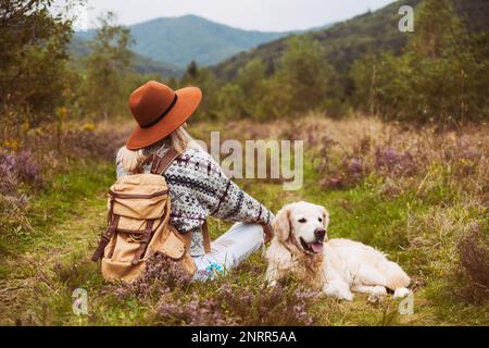 Young hippie blond hair woman in wool sweater with textile backpack and smiling happy golden retriever dog sitting on grass in summer mountains valley Stock Photo