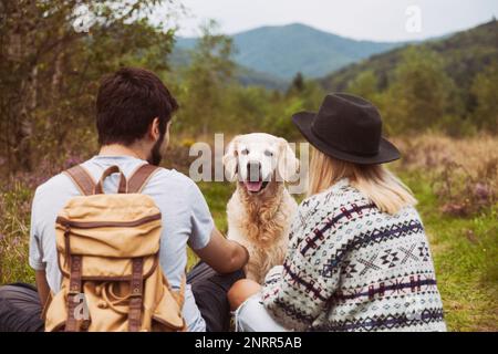 Young couple with smiling happy golden retriever dog sitting on grass in summer mountains valley. Pets travel, hiking and weekend activities concept. Stock Photo