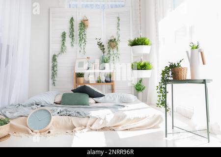 Light modern scandinavian bedroom interrior with bed, pillows, plaids, clock, shelves, green plants in baskets. Mock up morning pattern background. Stock Photo