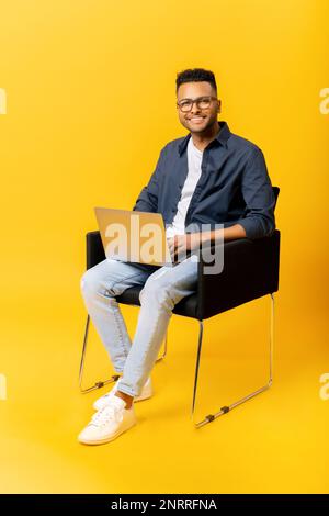 Cheerful indian guy using laptop in casual shirt and glasses using laptop sitting on the chair isolated on yellow, freelancer guy is working remotely, developer coding, student studying, full length Stock Photo