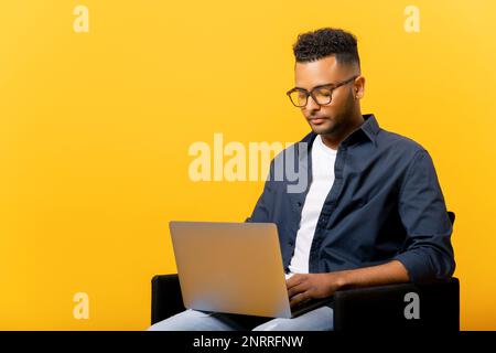 Concentrated indian guy using laptop in casual shirt and glasses using laptop sitting isolated on yellow background, freelancer guy is working remotely, developer coding, student studying on distance Stock Photo