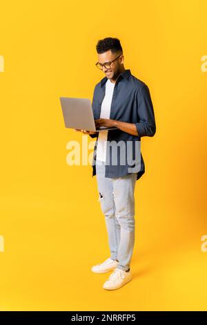 Full length photo of charismatic indian man is standing and typing on the laptop, handsome guy in casual wear working remotely, responding emails, copy space Stock Photo