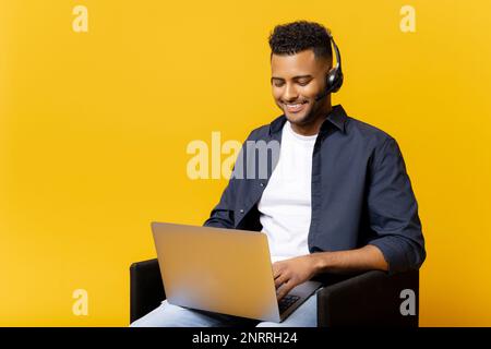 Concentrated indian man wearing headset holding virtual video conference sitting on the chair with a laptop computer on the laps, guy studying online, involved webinar Stock Photo