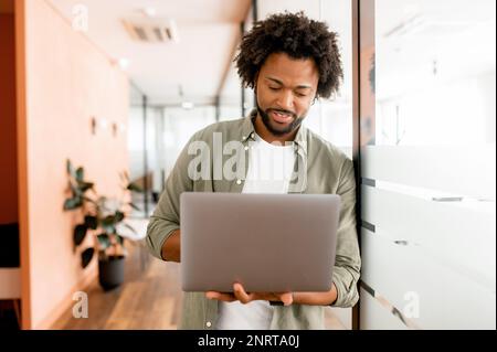 Serious concentrated determined young bearded office worker in casual clothes, stands in office hallway, typing on laptop, freelancer is working on project, writing email to client Stock Photo