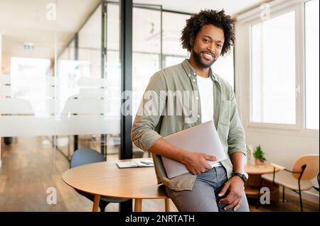 Portrait of confident African-American businessman standing with laptop, male office employee in smart casual wear looking at the camera and smiles, leaned back at te desk, ceo, manager, sturtup owner Stock Photo