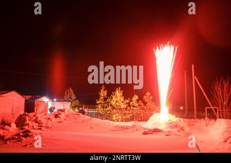 Bright flashes of fireworks in a garden plot in Siberia in winter with Christmas trees decorated with garlands on the snow at night. Stock Photo