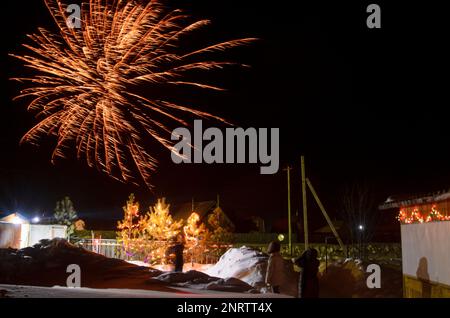 Bright flashes of fireworks in a garden plot in Siberia in winter with Christmas trees decorated with garlands on the snow at night. Stock Photo