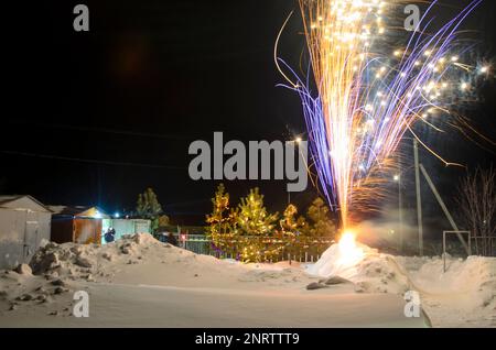 Bright flashes of fireworks in a garden plot in Siberia in winter with Christmas trees decorated with garlands on the snow at night. Stock Photo