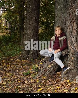 A handsome guy sits on the root of a huge oak tree leaning against the trunk of a tree and smiles Stock Photo