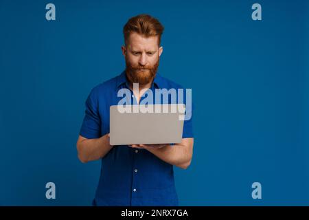 Adult handsome stylish redhead bearded man holding and using laptop , while standing over isolated blue background Stock Photo