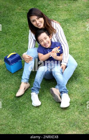 Latino mom and 6 year old boy sitting on the grass with lunch box and colors for back to school Stock Photo